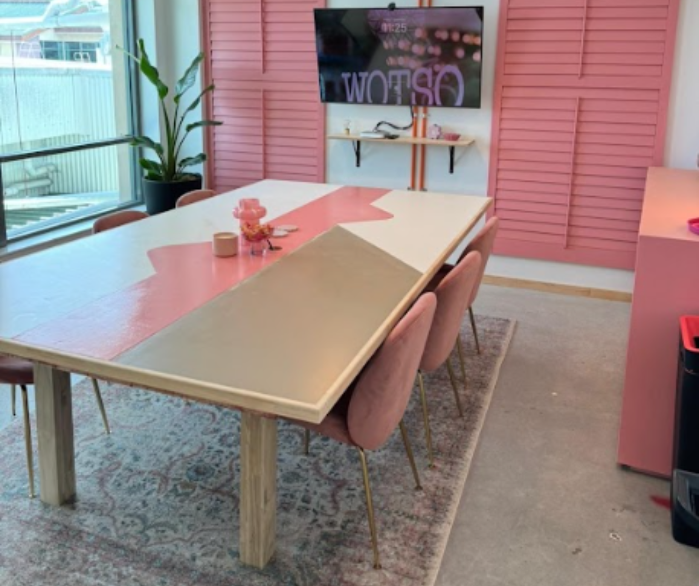 Pink-accented conference room with long table, pink chairs, and a wall poster near a window. Pink-accented conference room with long table, pink chairs, and a wall poster near a window.