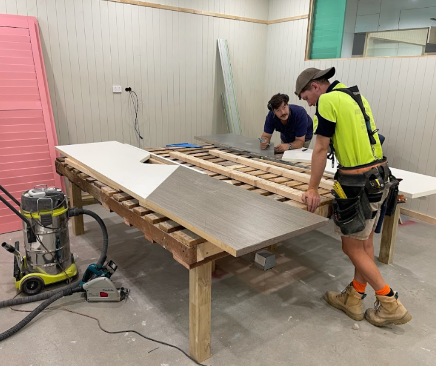 Two workers installing wood panels on a large tabletop in a workshop, with tools nearby. Two workers installing wood panels on a large tabletop in a workshop, with tools nearby.