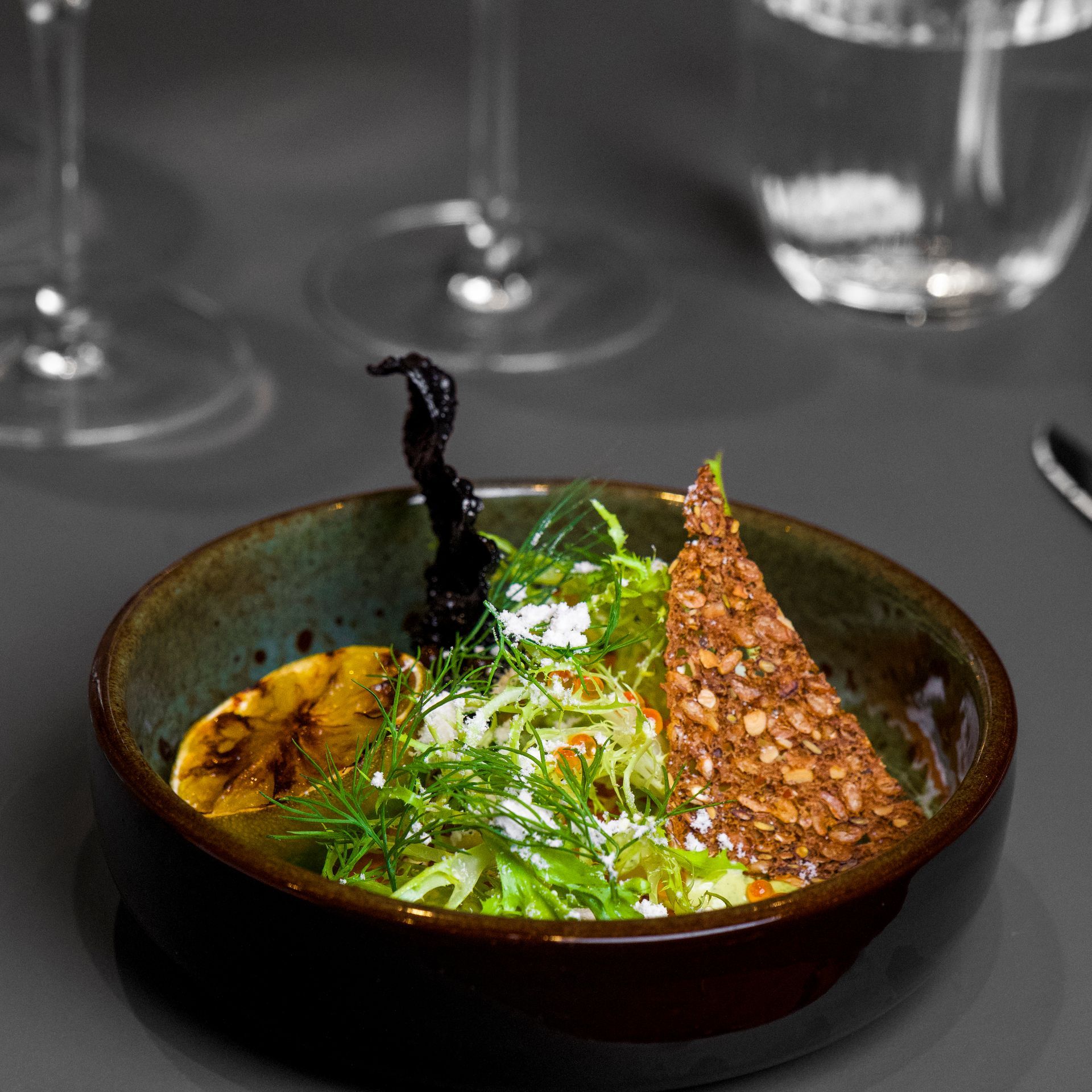 En skål salat med grillet citron, krydderurter og en kiks på et mørkegråt bord.