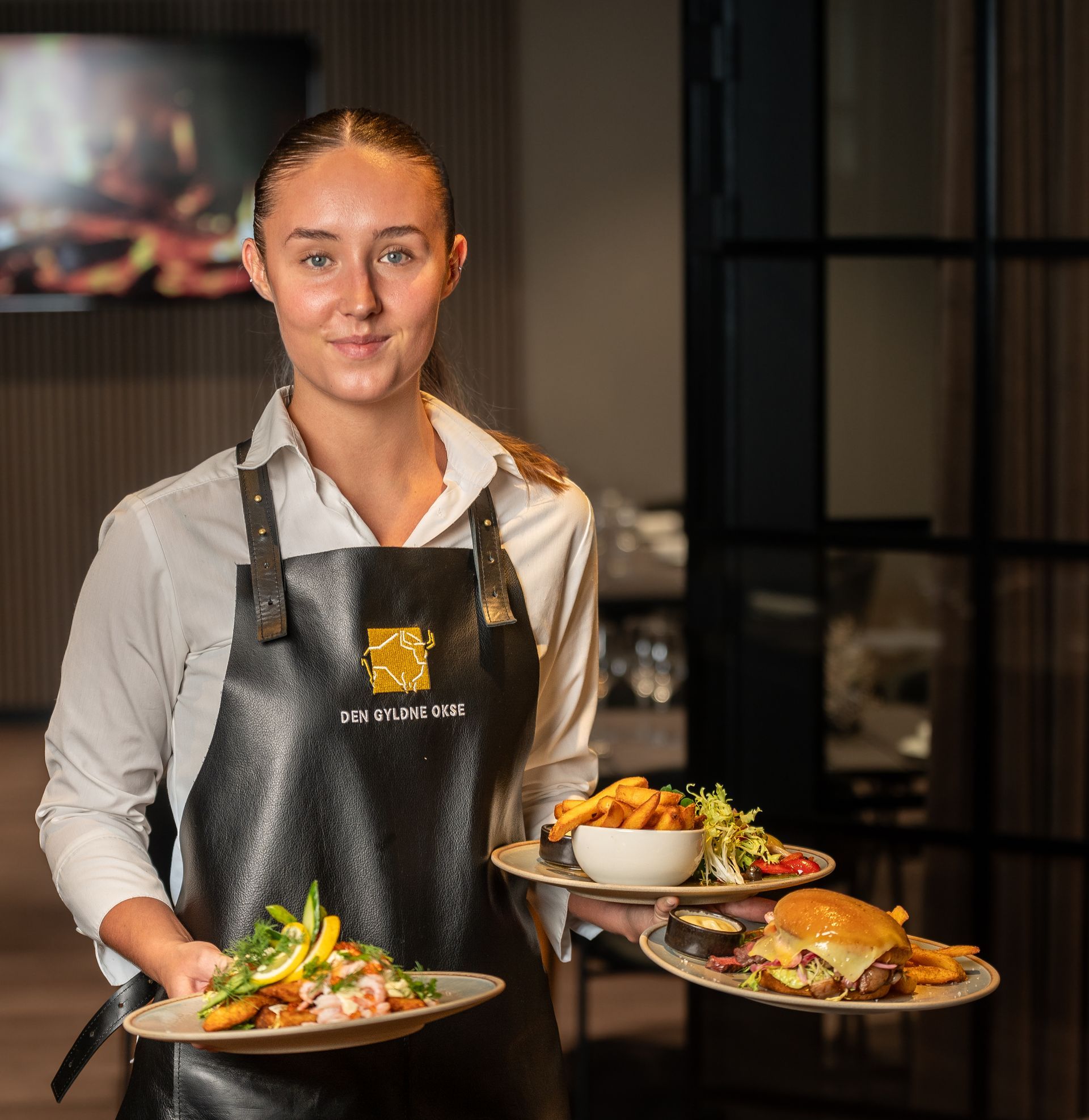 Tjener holder tallerkener med mad: fisk, burger og pommes frites. Restaurantinteriør.