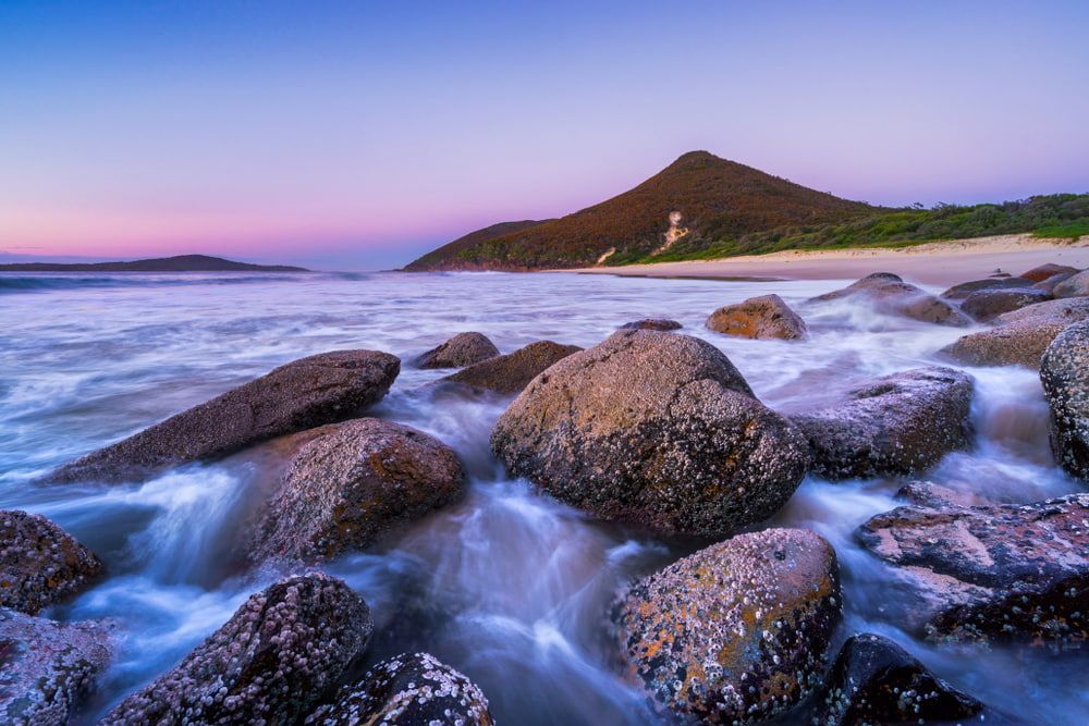 A Rocky Beach with A Mountain in The Background and Waves Crashing on The Rocks — Concrete Colour Solutions in Port Stephens, NSW