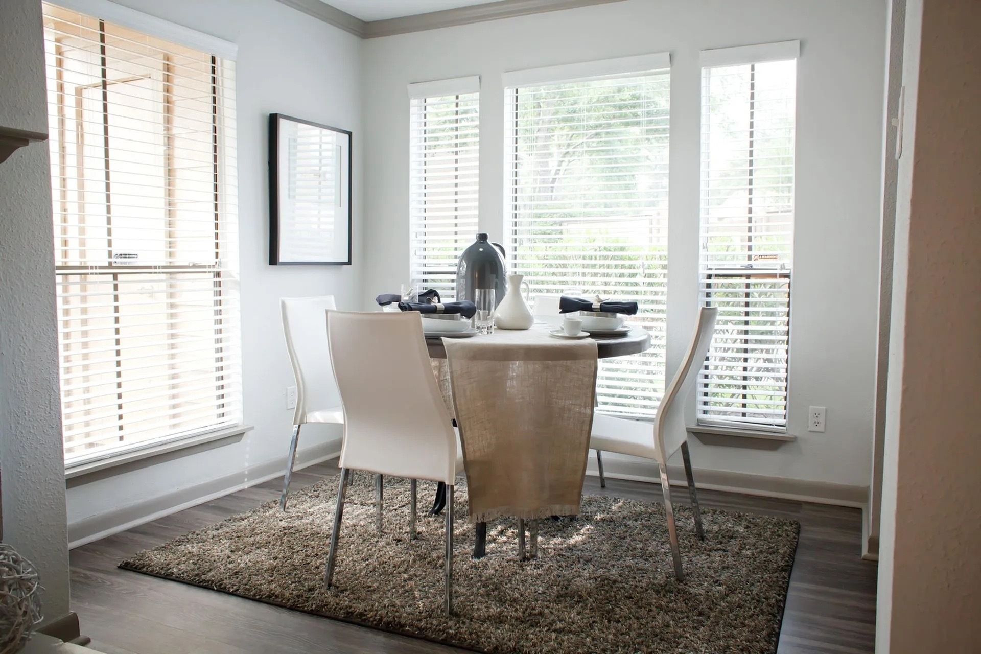 Dining area in a modern apartment with a round table, white chairs, and large sunlit windows.