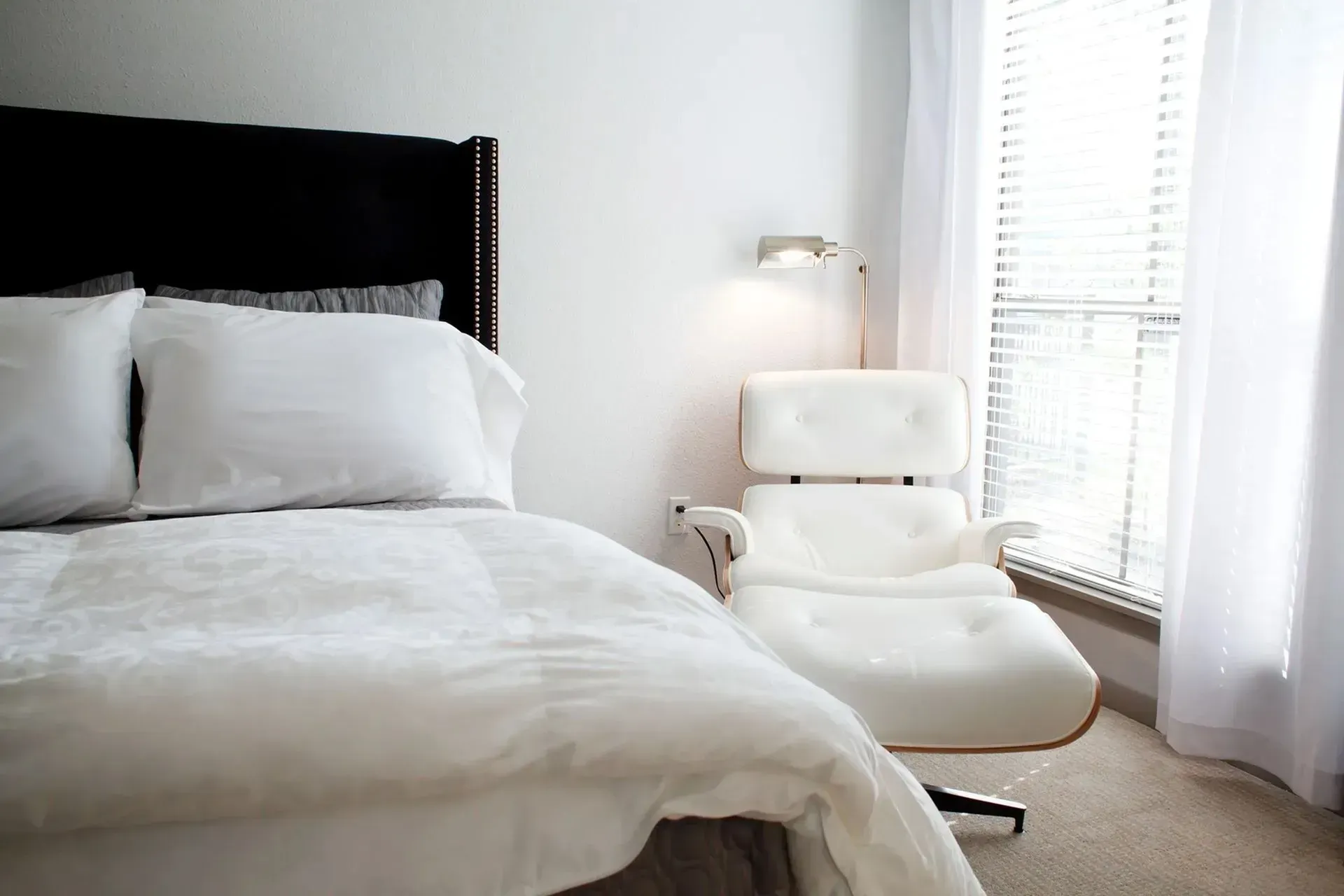 White bedroom with bed and lounge chair near window with blinds and sheer curtains.