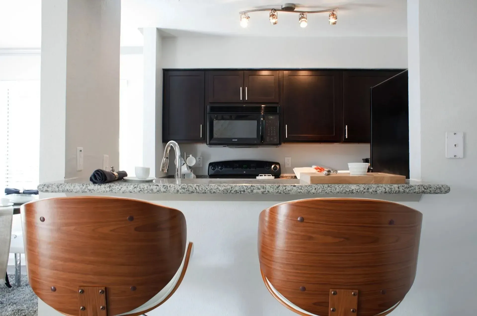 Two wooden barstools face a kitchen countertop. Dark cabinets, microwave, and under-cabinet lighting are visible.