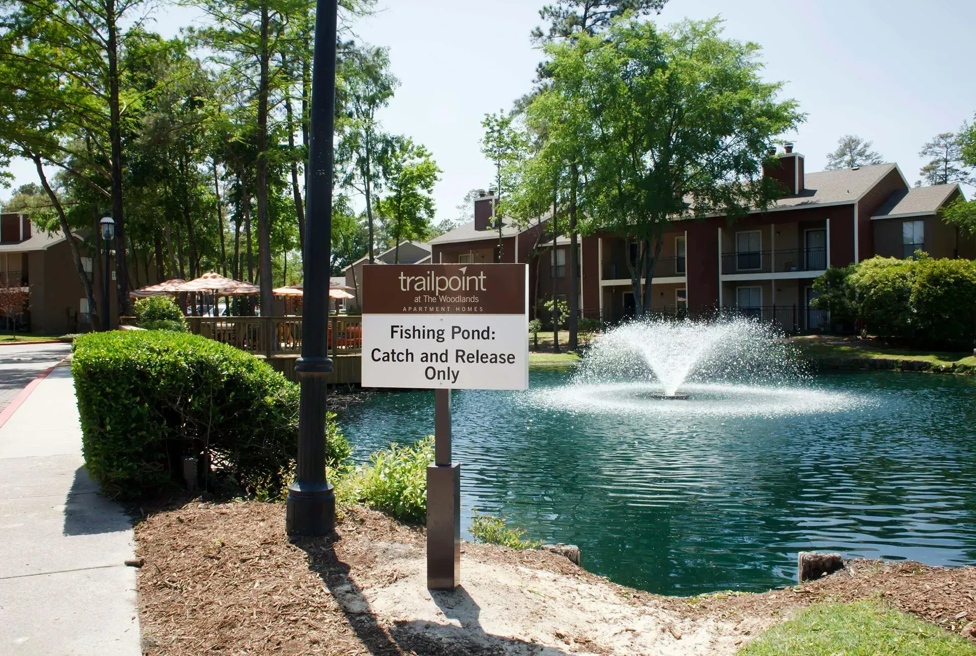 A sign for a fishing pond with a fountain in front of a brown brick building, surrounded by trees.