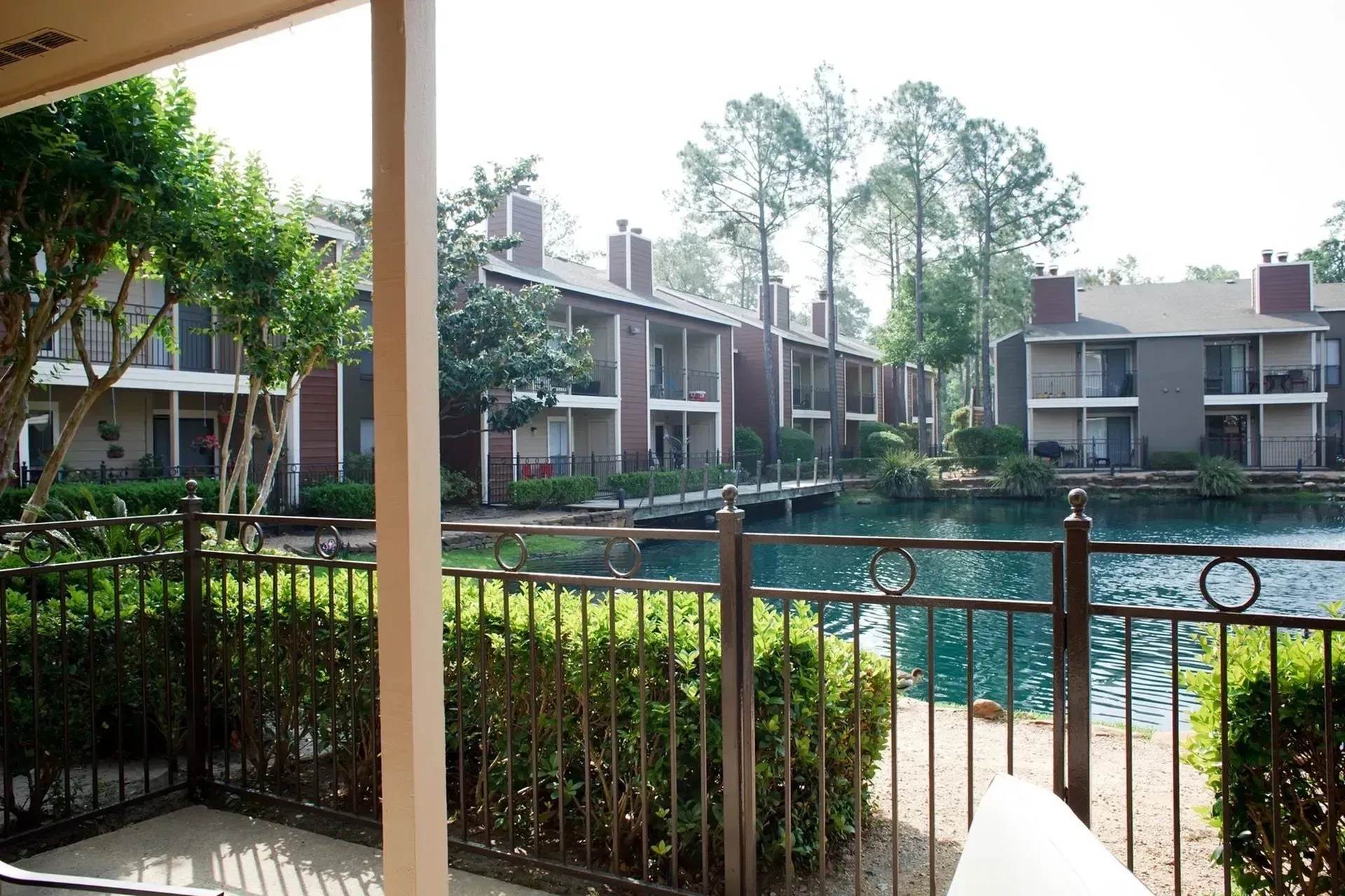 Patio overlooking a pond with apartment buildings in the background and a black fence.
