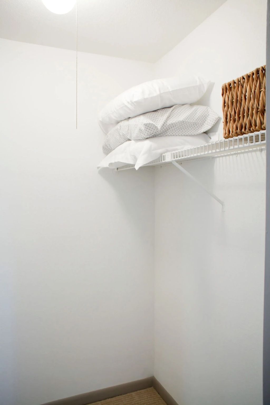 Small white closet with a wire shelf, stacked pillows, and a woven basket.