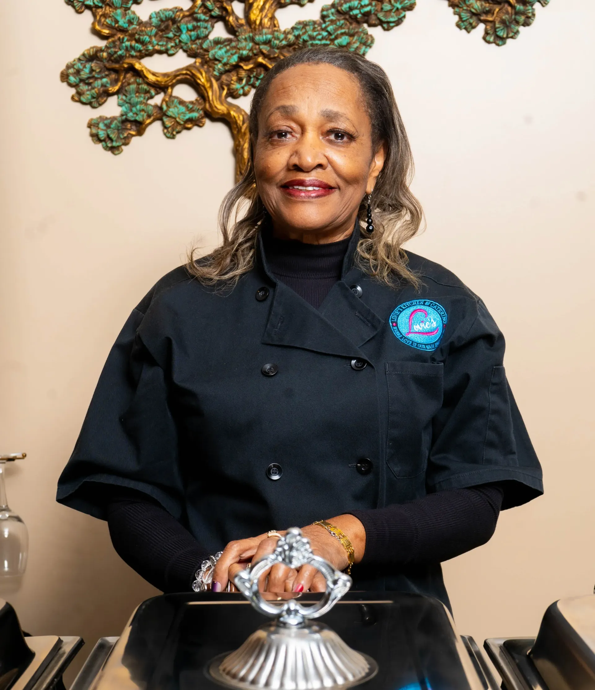 Woman in a black chef coat standing behind a silver cloche, smiling in a kitchen.