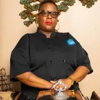 Chef in a black uniform with glasses, seated at a table with hands clasped, in a decorated indoor setting.