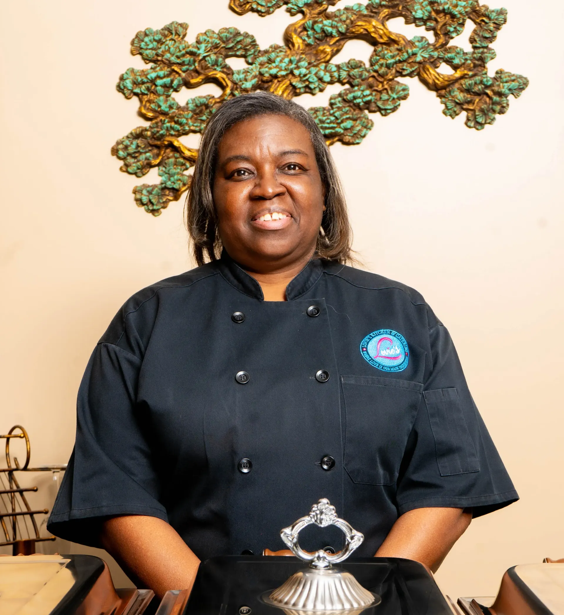 Smiling chef in black uniform standing behind a counter with a decorative tree sculpture in the background