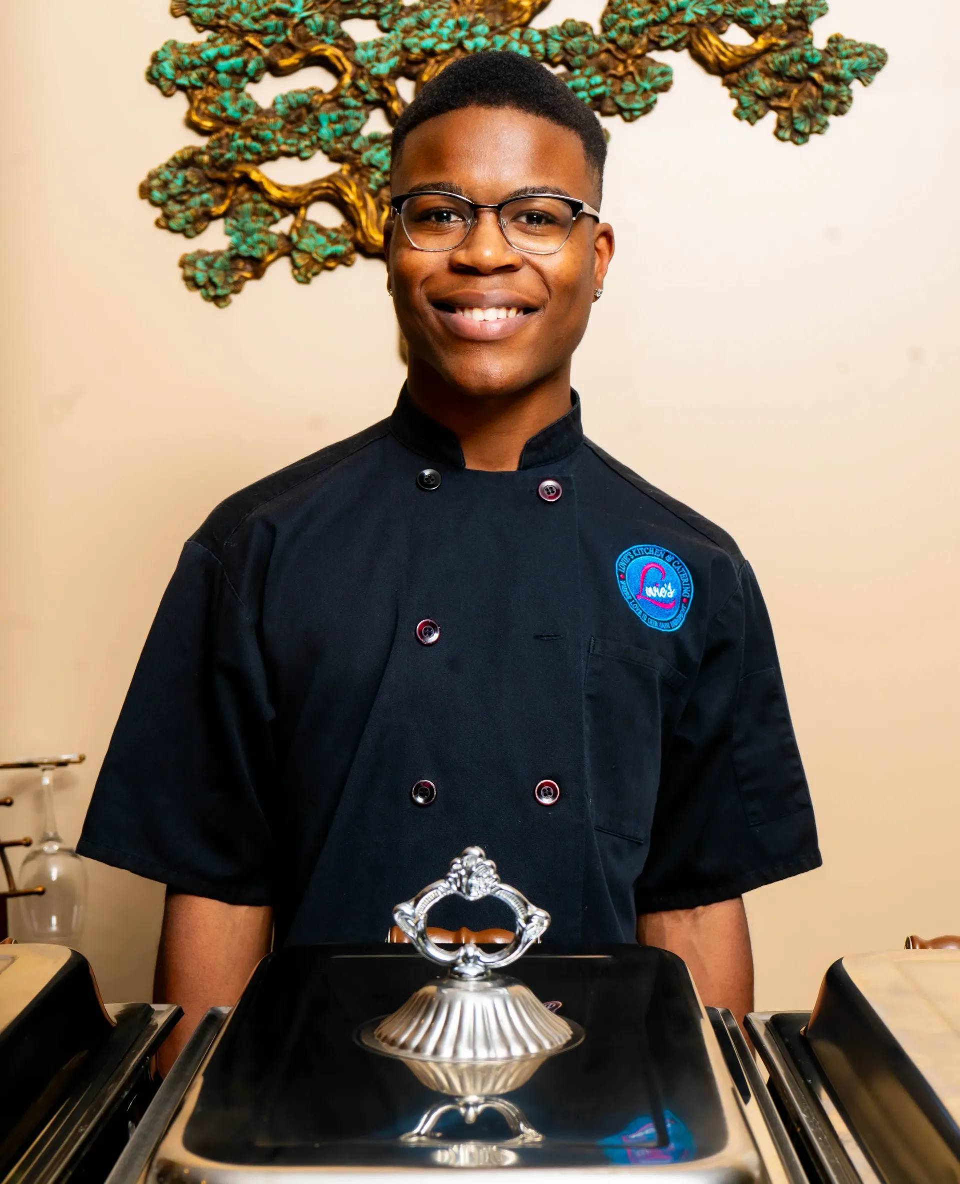 Smiling chef in black uniform standing behind a silver chafing dish in a restaurant setting
