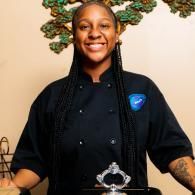Smiling chef in a black uniform standing behind a counter with a decorative wall accent behind them