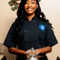 Person in a black chef uniform smiling and holding a glass serving dish indoors