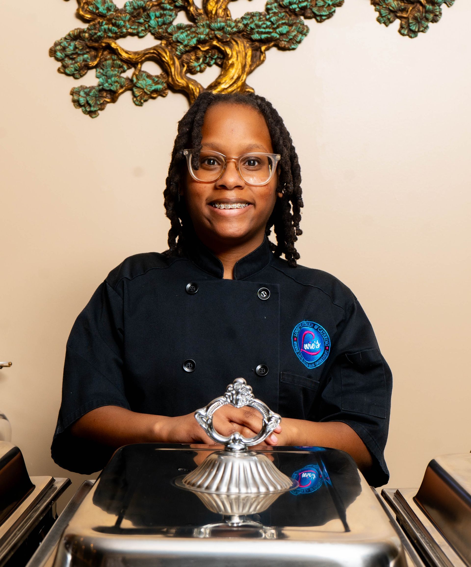 Smiling person in black chef coat holding a dessert plate at a counter
