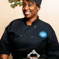 Chef in black uniform smiling, wearing a black hat and holding a trophy.