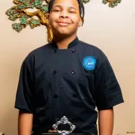 Person in a black chef coat standing indoors with a blue badge and a decorative wall behind them