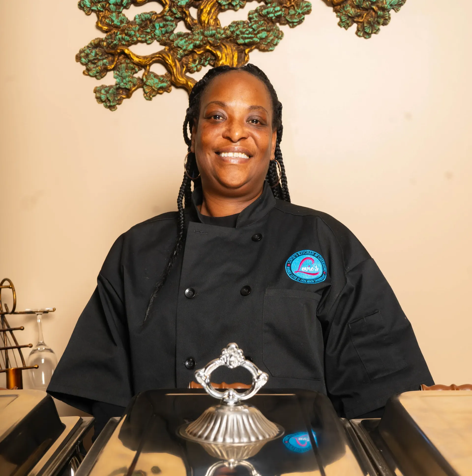 Chef in black uniform smiling behind a kitchen counter with a decorative dish cover.