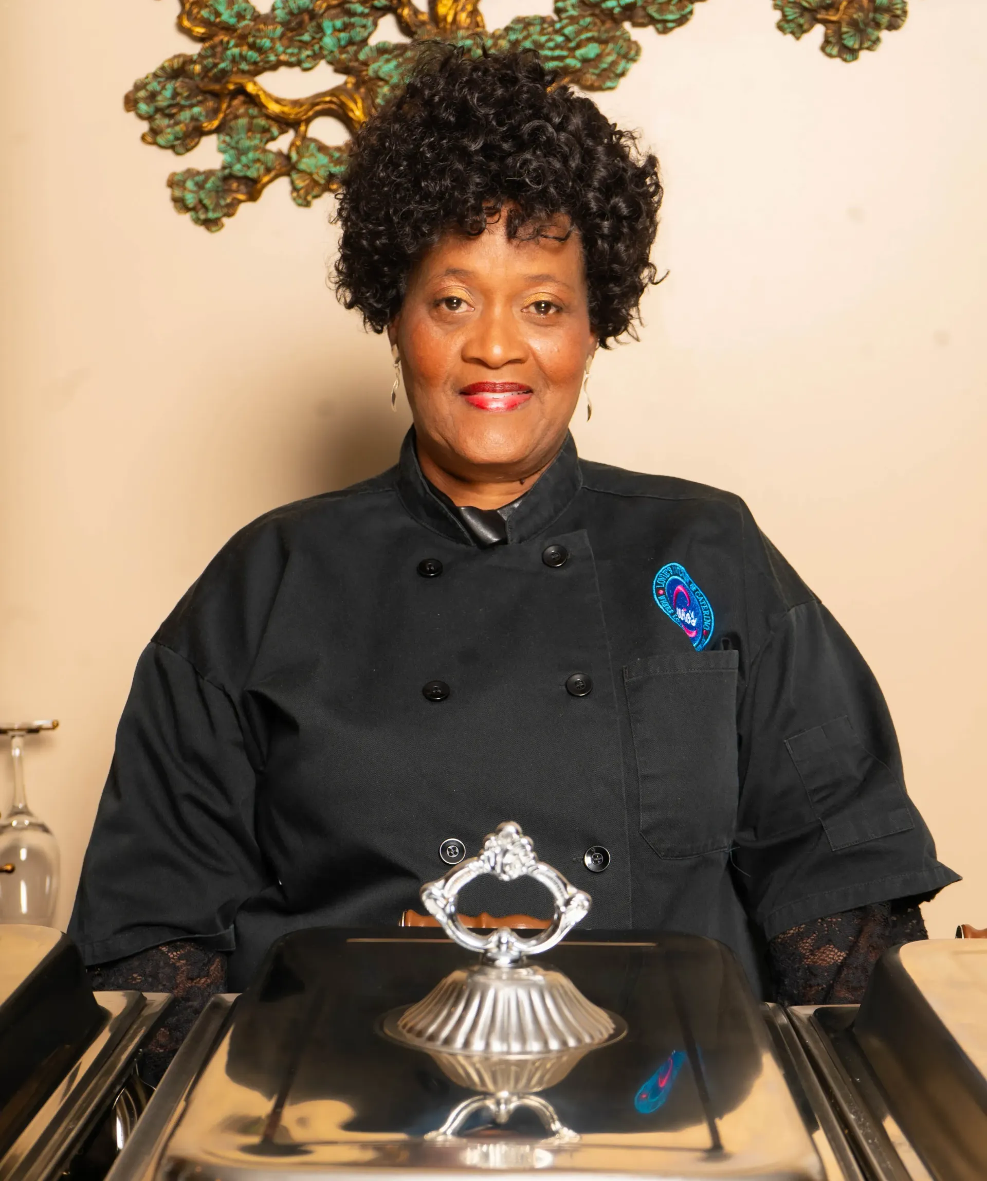 Chef in black uniform standing behind a silver dessert display, smiling in a formal dining setting.