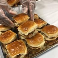 Gloved hands stacking small sliders on a tray, with several golden-brown buns arranged in rows.