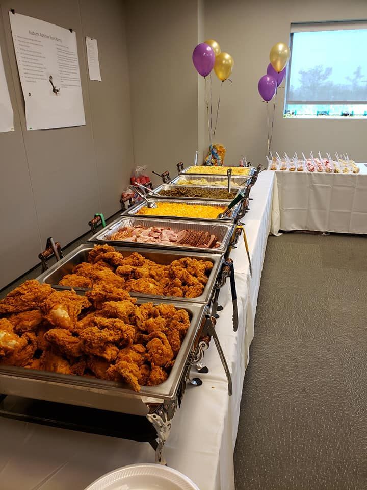 Buffet table with fried chicken, side dishes, and purple-gold balloons in a decorated room
