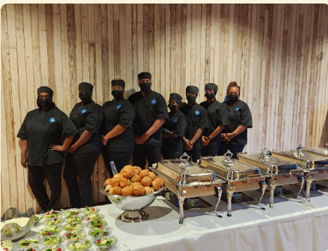 Caterers in black uniforms stand behind a buffet table with trays and platters of food