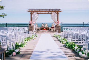 Outdoor wedding ceremony setup with white chairs, floral aisle, and pergola altar by the water