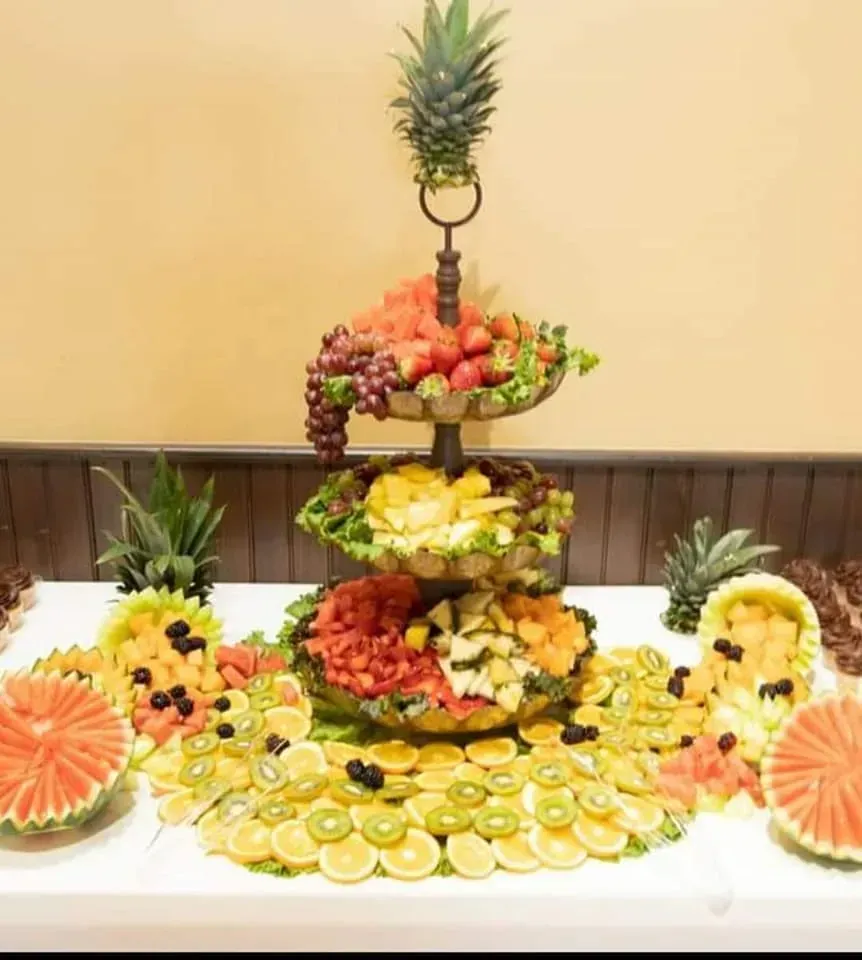 Fruit and vegetable display arranged in tiers, topped with a pineapple and surrounded by sliced citrus and melons.
