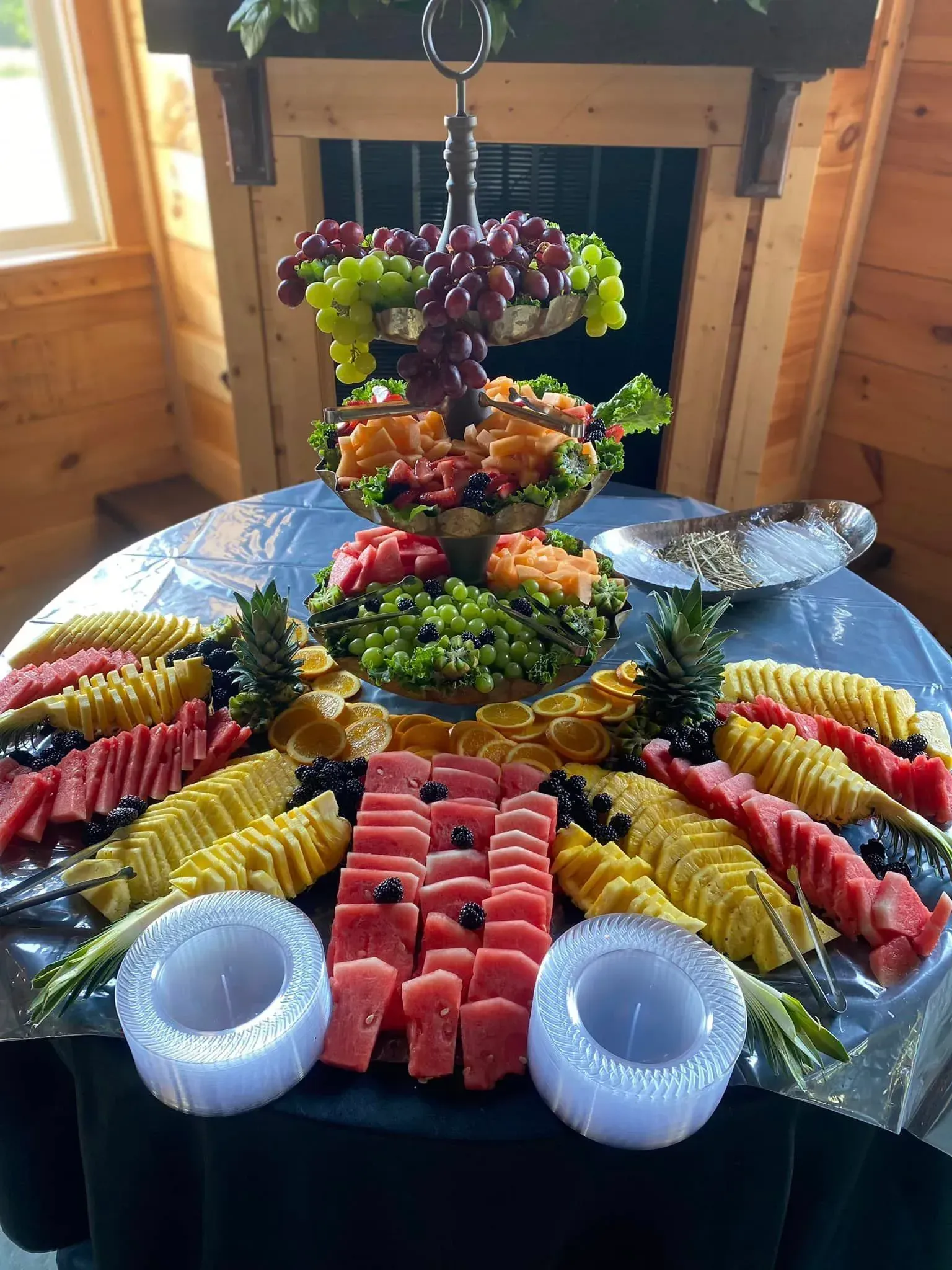 Fruit platter display on a table, arranged as a colorful fan with bananas, pineapple, grapes, kiwi, and melon in a wood room
