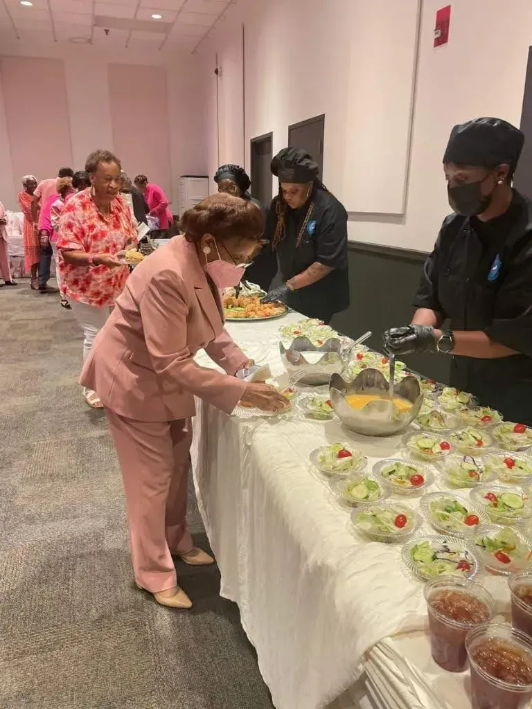 People serving food at a buffet table with salads and desserts in a bright hallway