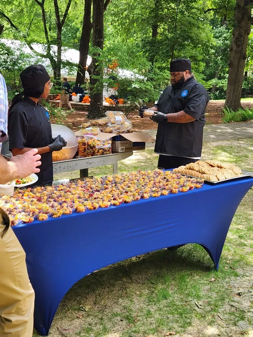 People serving food at an outdoor buffet table with a blue tablecloth in a wooded park