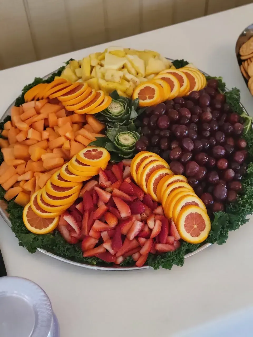 Fruit platter with neatly arranged sliced oranges, grapes, strawberries, melon, and pineapple on a white table