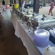 Buffet table with covered serving trays and a large water dispenser at an outdoor event