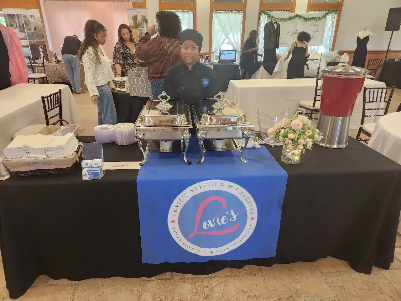 Catered event table with a blue banner, food trays, and floral centerpiece in a banquet hall