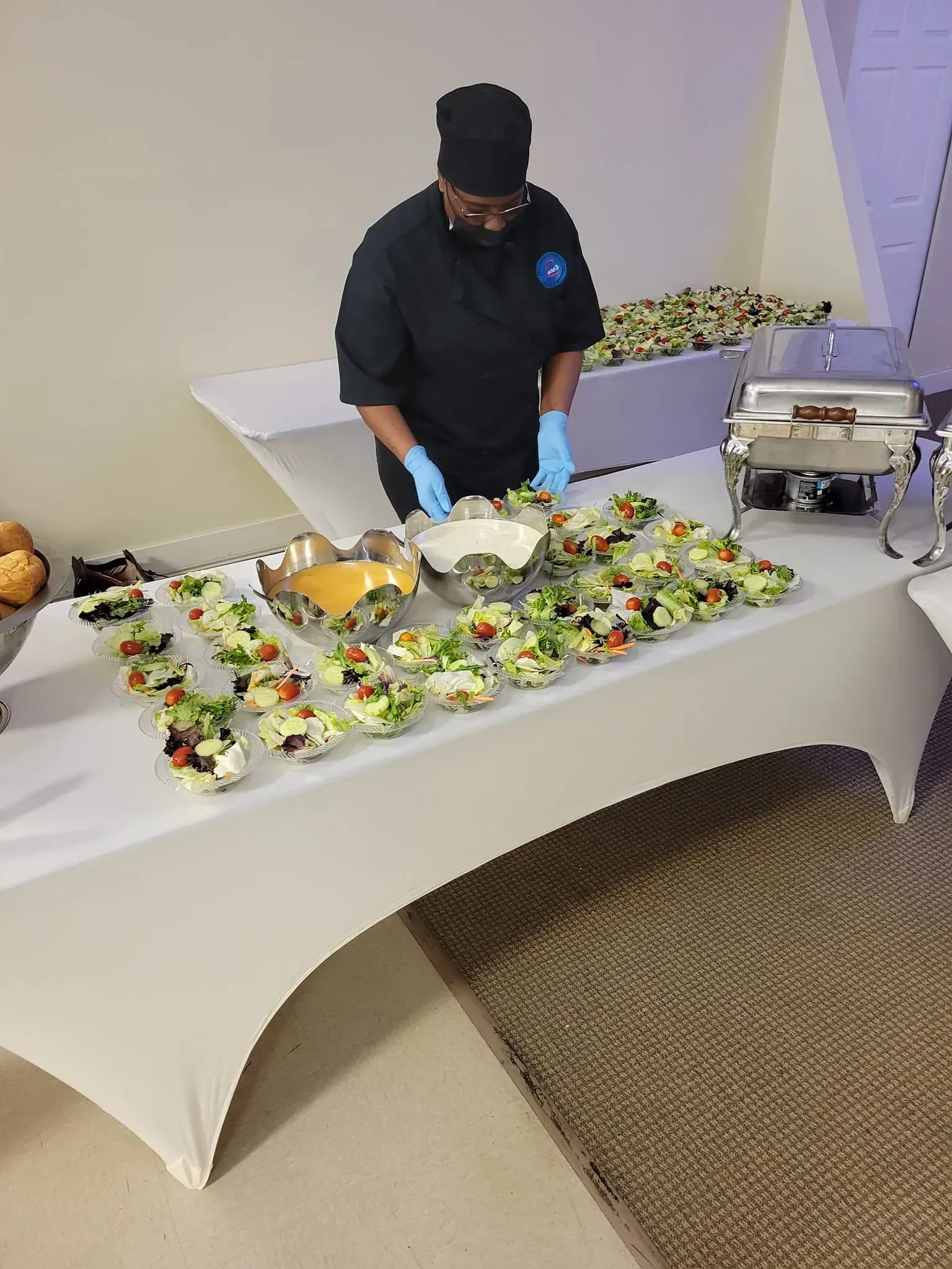 Caterer in black gloves and mask serves a buffet table with salad and sandwiches at an indoor event.