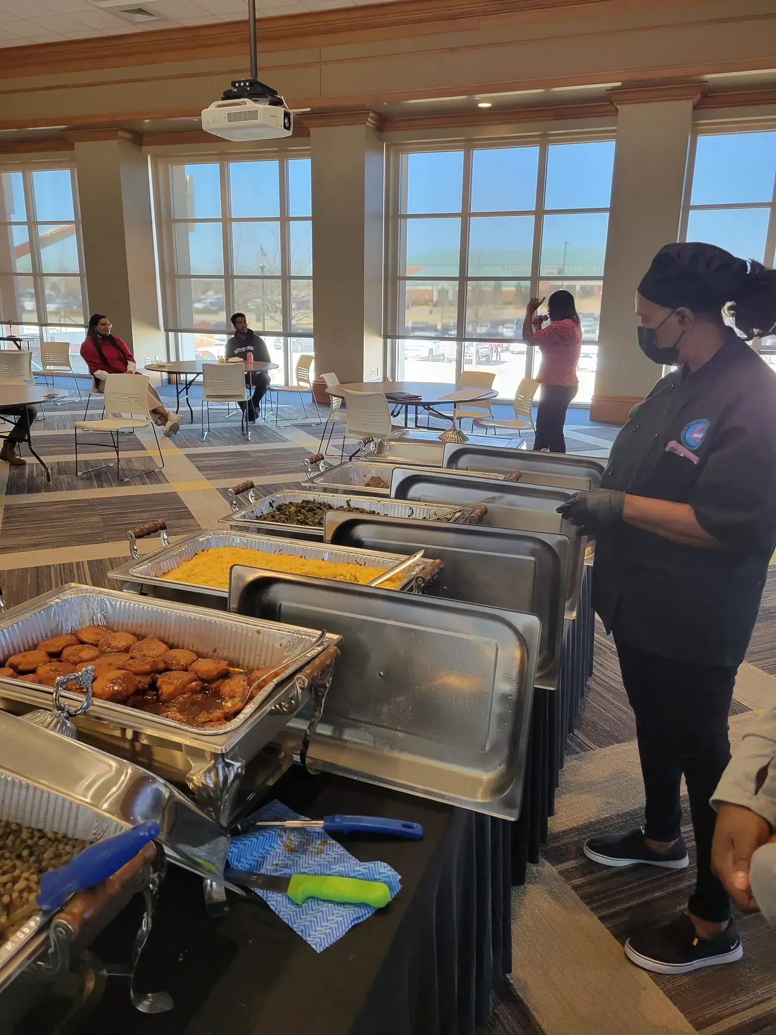 Buffet line with chafing dishes in a bright room, with people serving food near large windows.