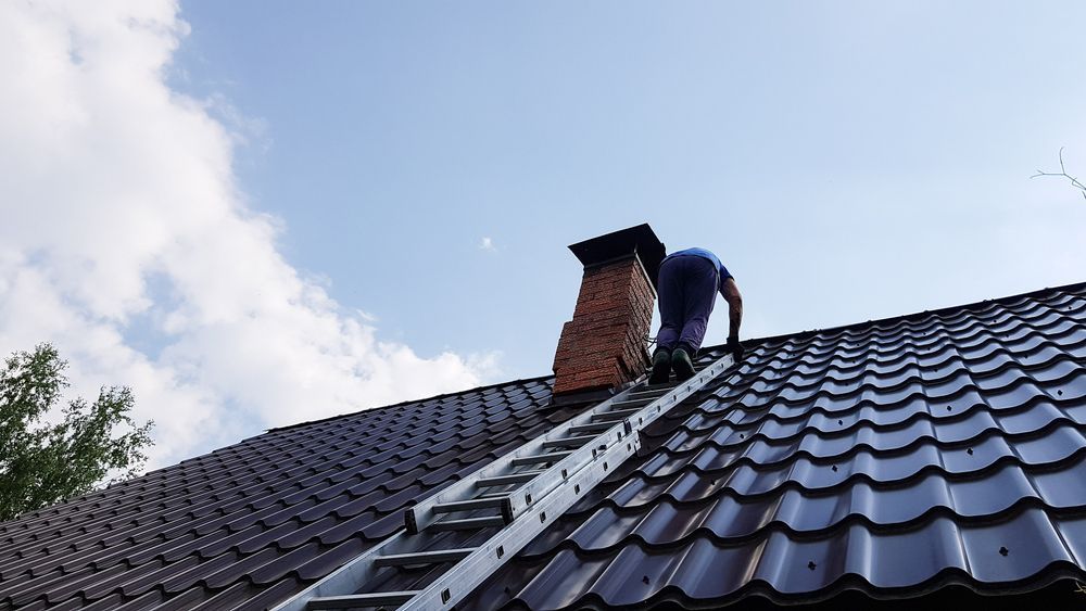 A Person on a Ladder Repairs a Brick Chimney on a Dark Tiled Roof — The Flue Pro In Huntingfield, TAS
