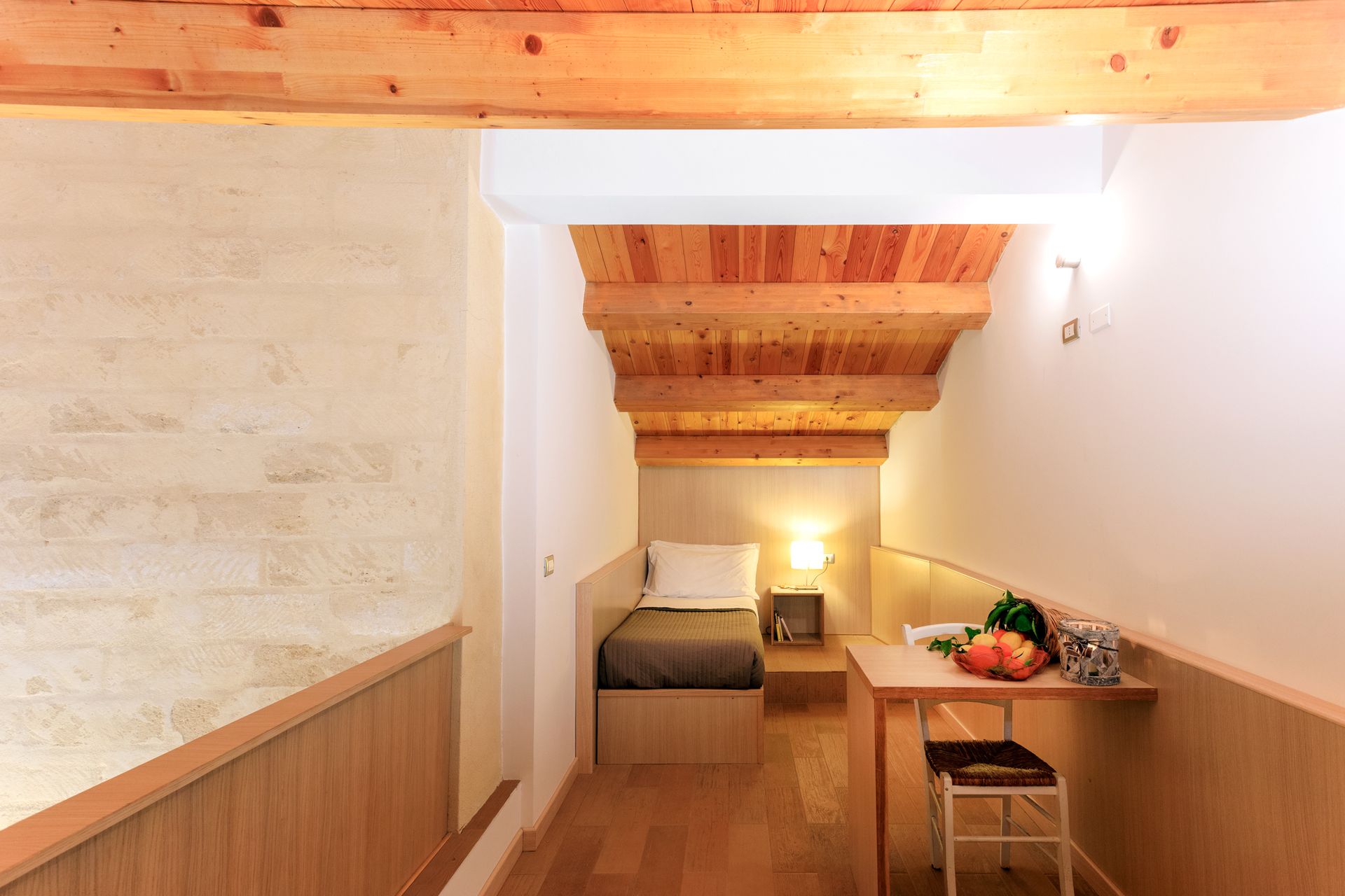 Camera da letto stretta in legno con letto, scrivania e luce calda sotto le travi in legno a vista.