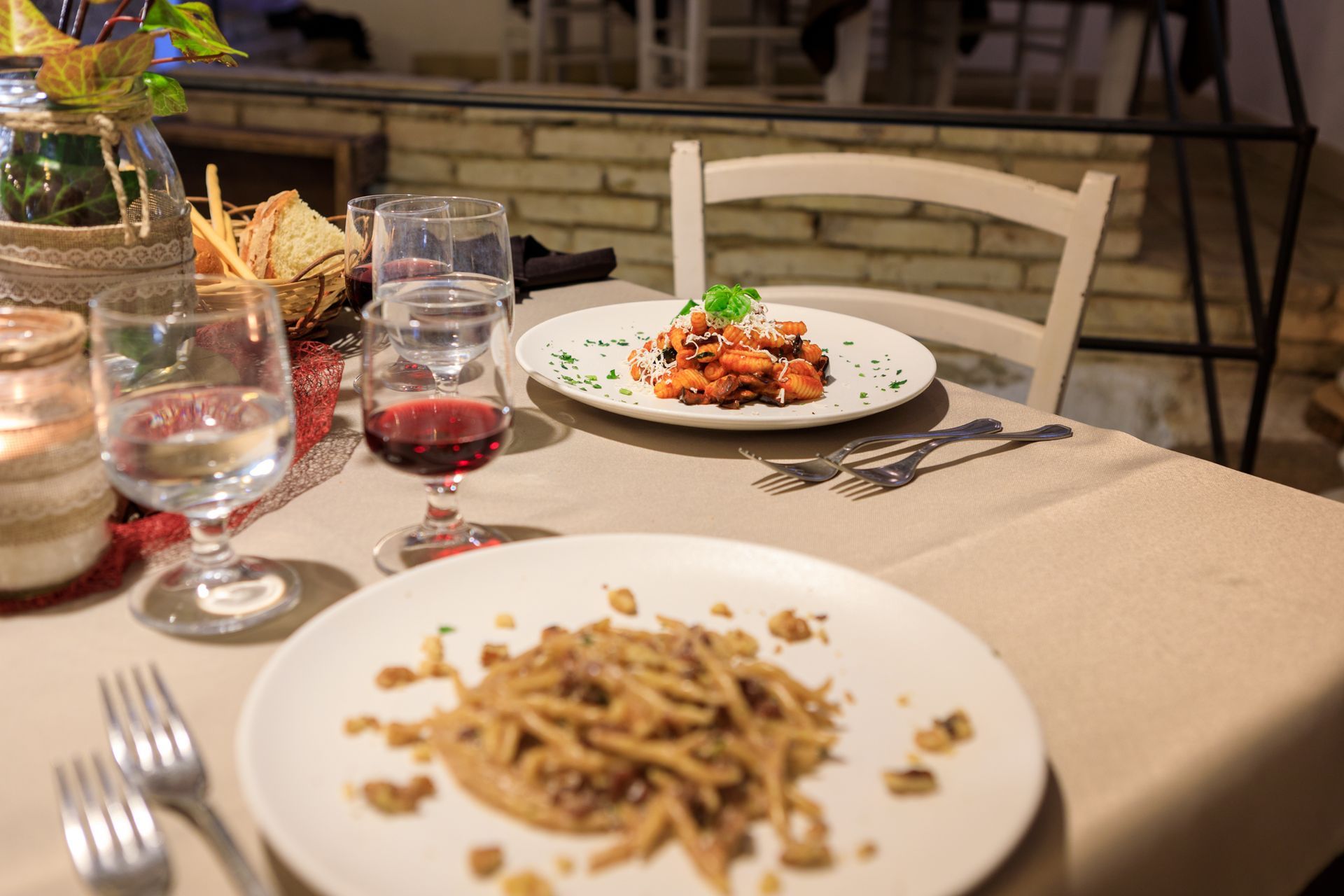 Tavolo da ristorante apparecchiato con pasta, bicchieri da vino e un piatto su una tovaglia beige.
