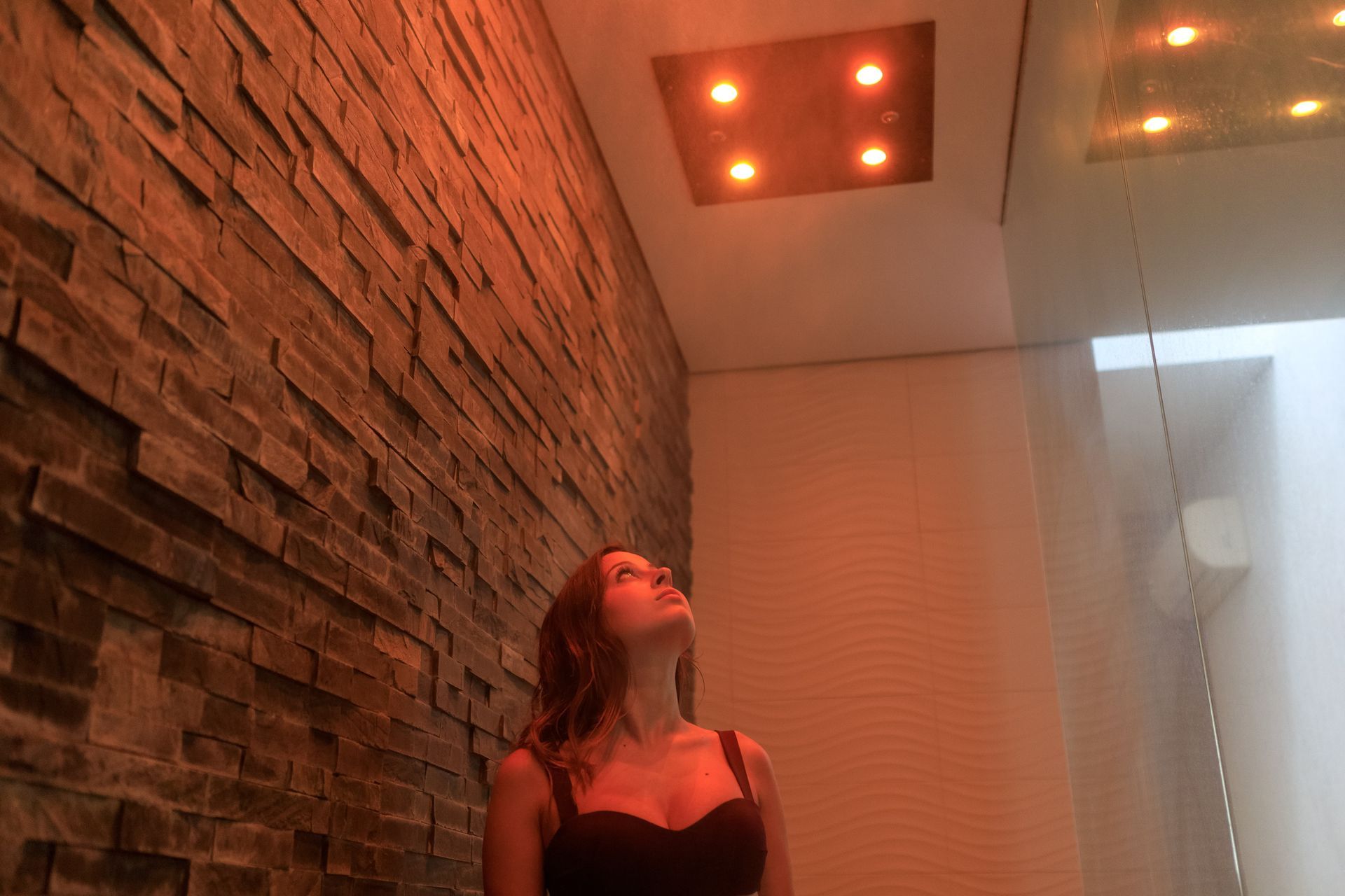 Persona che guarda in alto in un corridoio poco illuminato con muro di mattoni, luci arancioni sul soffitto e tende bianche