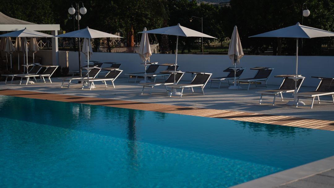 Piscina all'aperto con lettini, ombrelloni e un piccolo edificio sullo sfondo.