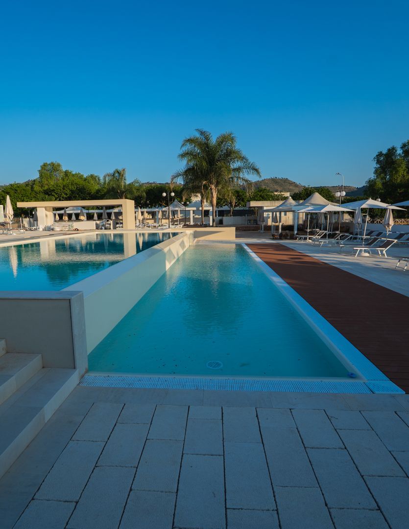 Piscina del resort con acqua turchese, lettini, palme e un cielo azzurro limpido al tramonto.