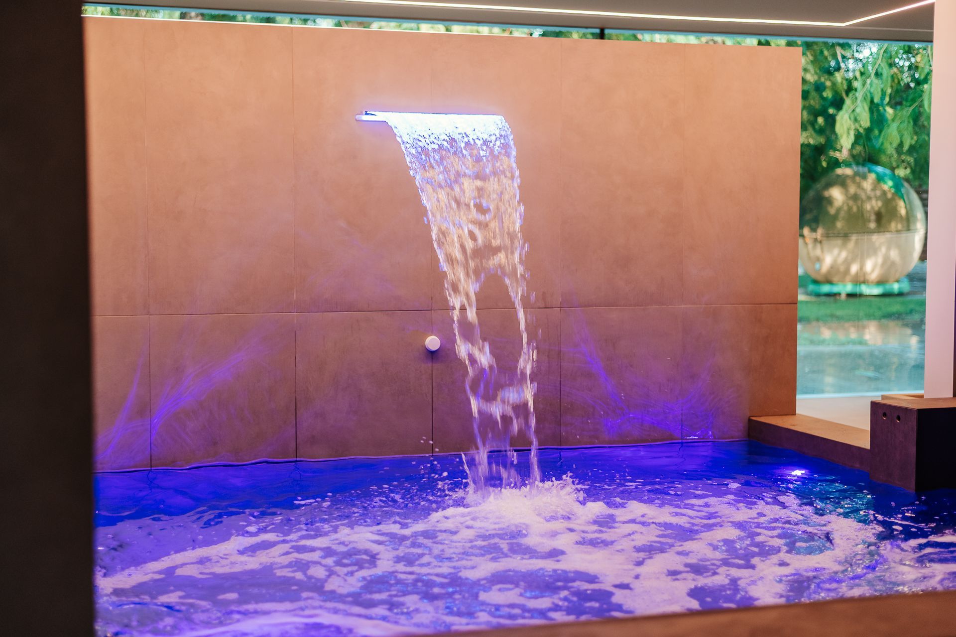 Piscina coperta con ampie vetrate, terrazza in legno e sdraio verde.