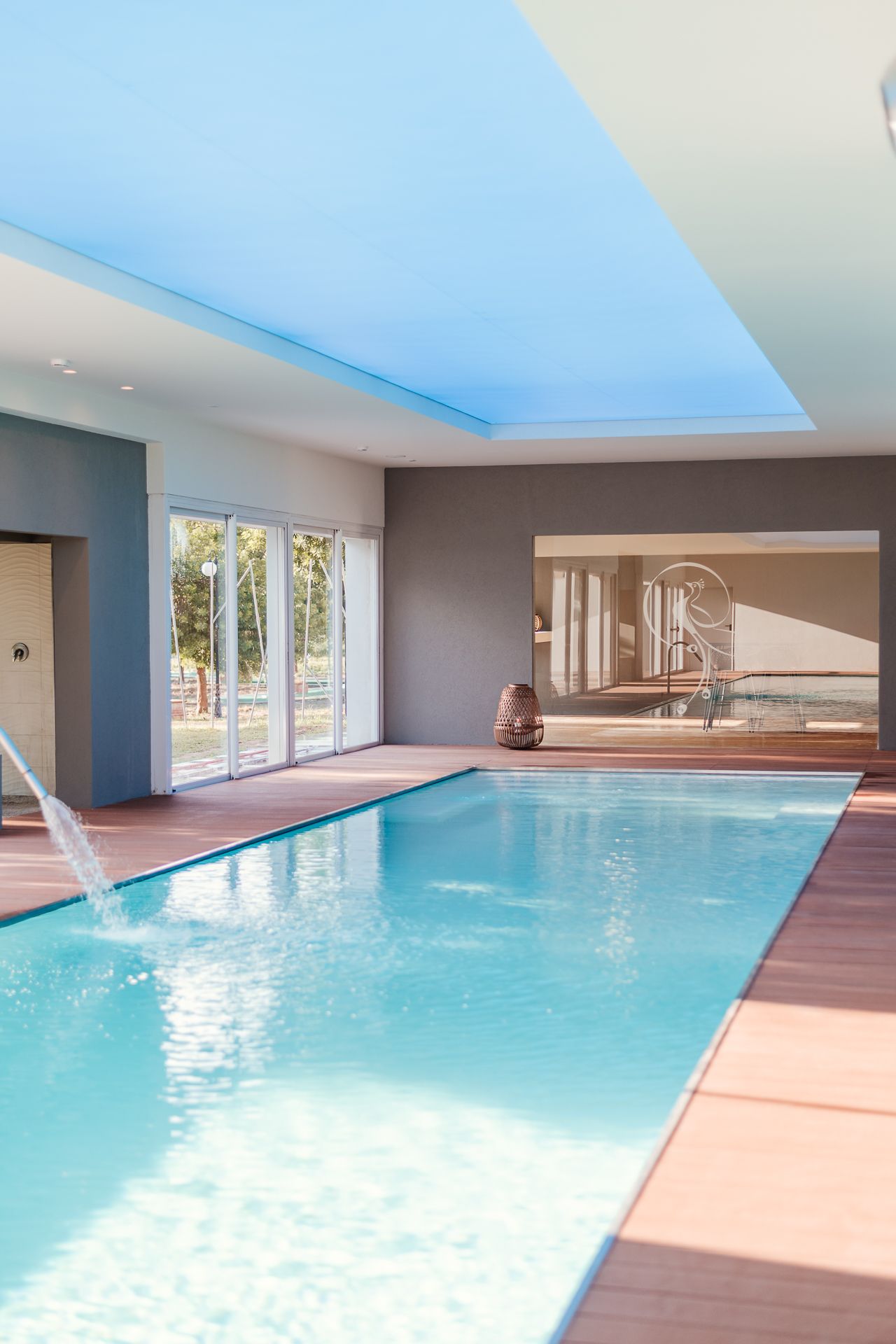 Piscina coperta con acqua turchese, terrazza in legno e lettini prendisole accanto a grandi finestre.