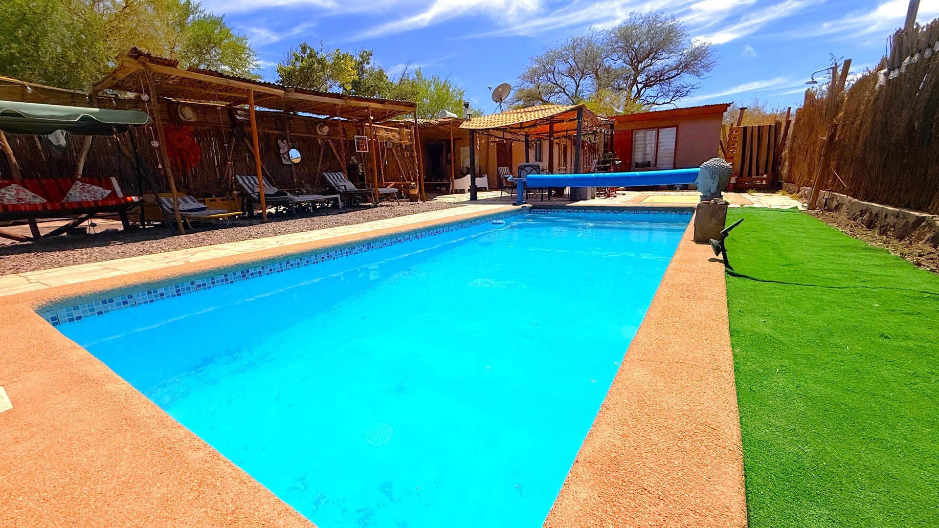 Piscina azul brillante en el patio trasero con terraza, asientos a la sombra y césped verde bajo un cielo soleado.