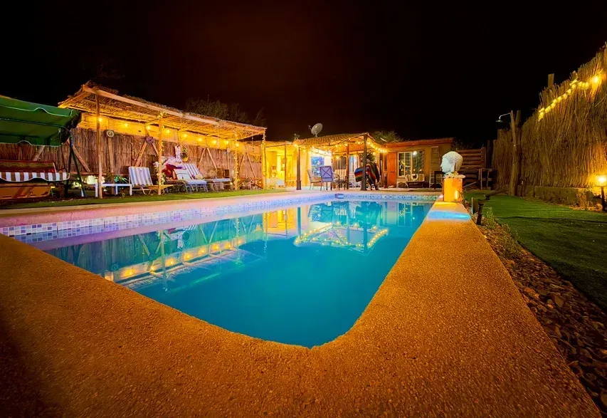 Una piscina exterior iluminada por la noche, rodeada de estructuras con techos de paja y luces cálidas de guirnalda.