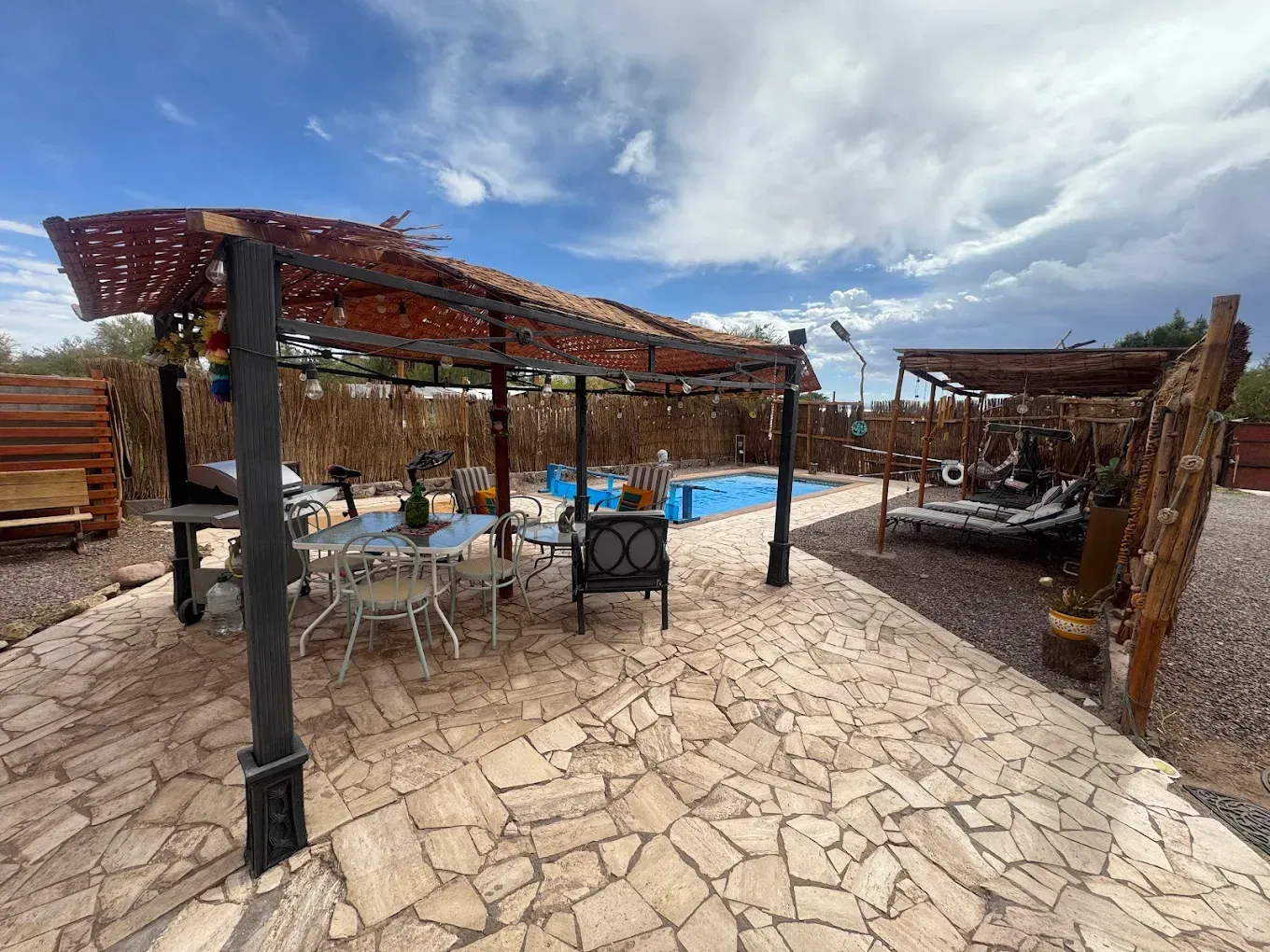 Un patio al aire libre con pérgola, mesa de comedor y asientos junto a una piscina bajo un cielo azul con nubes.