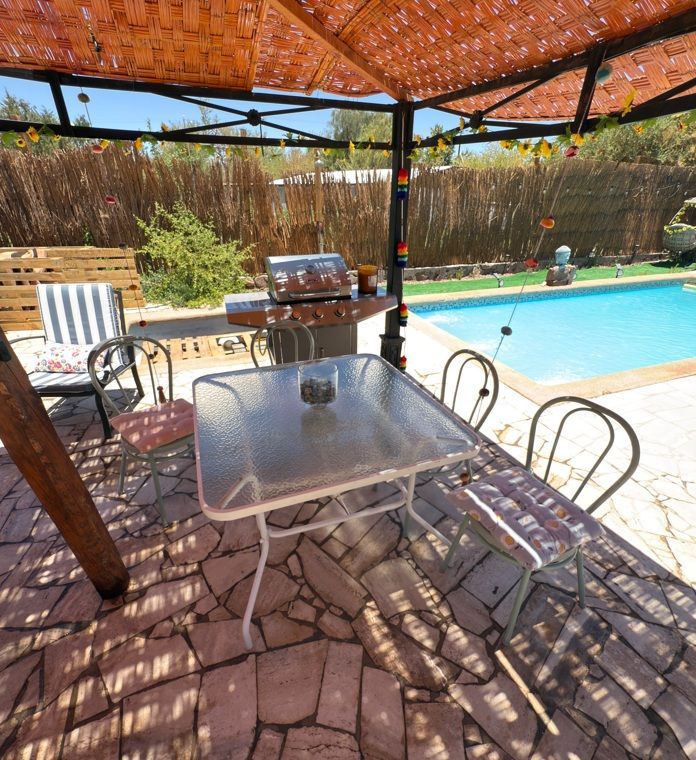 Zona de estar en el patio bajo una pérgola junto a una piscina en el jardín trasero, con una mesa de cristal y sillas.