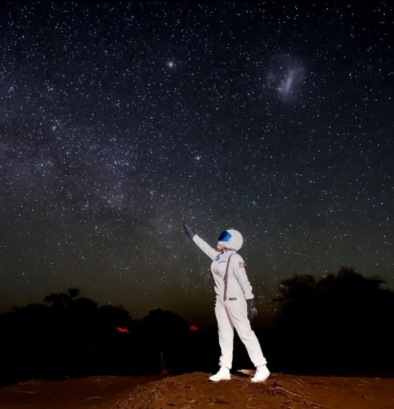 Astronauta con traje blanco señalando hacia un cielo nocturno estrellado.