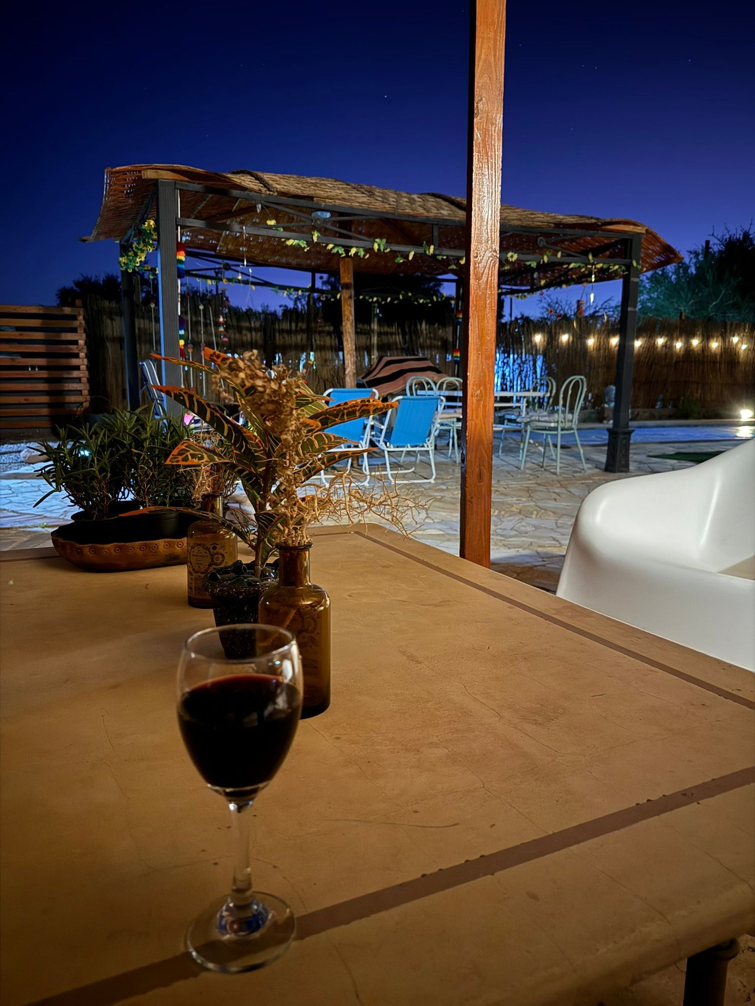 Una copa de vino sobre una mesa con vistas a una piscina iluminada y una pérgola al atardecer.