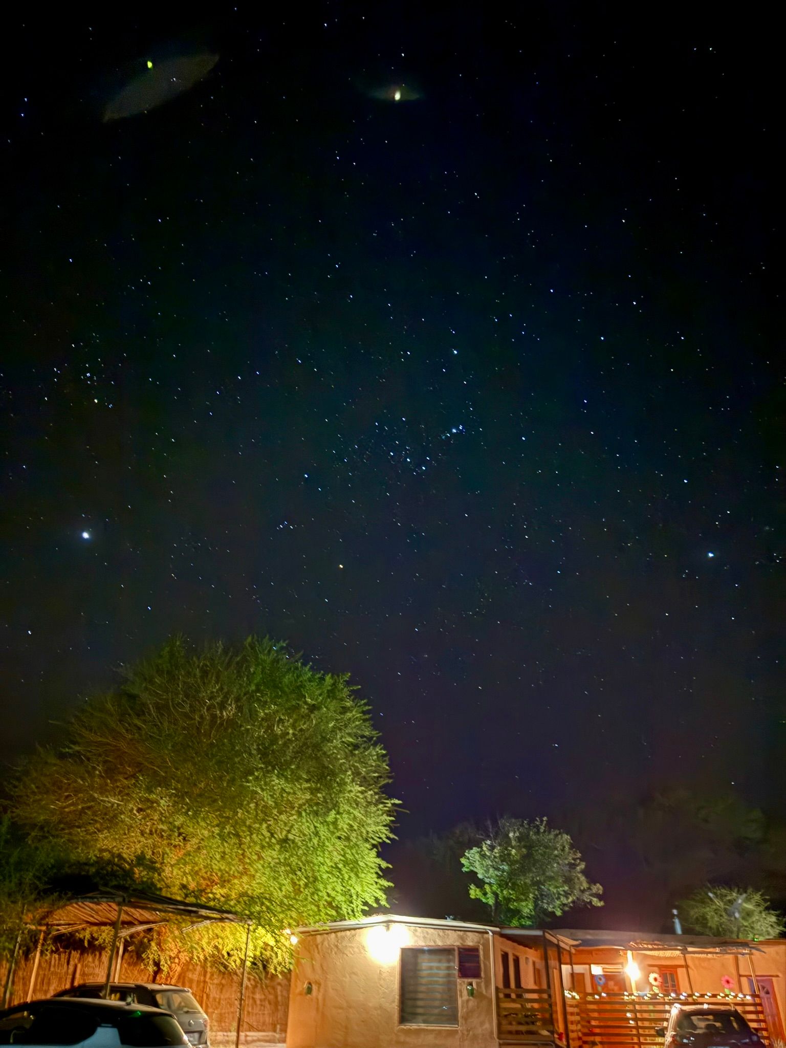 Cielo nocturno sobre un patio rústico con árboles y cabañas iluminadas por luces cálidas.
